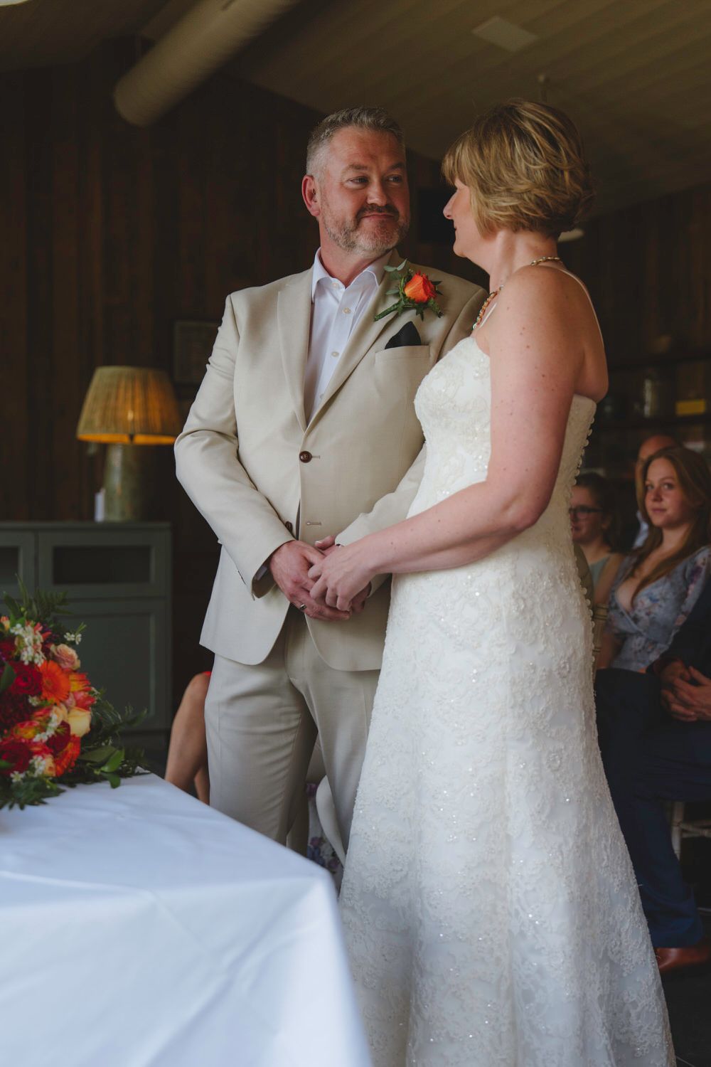 Bride and groom holding hands at indoor wedding ceremony, groom in beige suit with orange boutonniere.