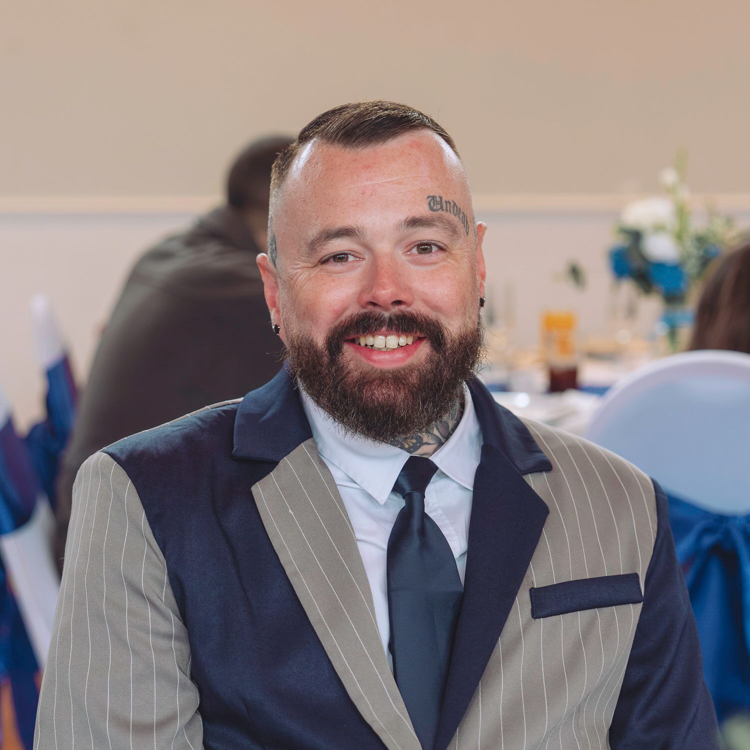 Smiling bearded man with tattoos wearing a stylish suit and tie at a formal event.