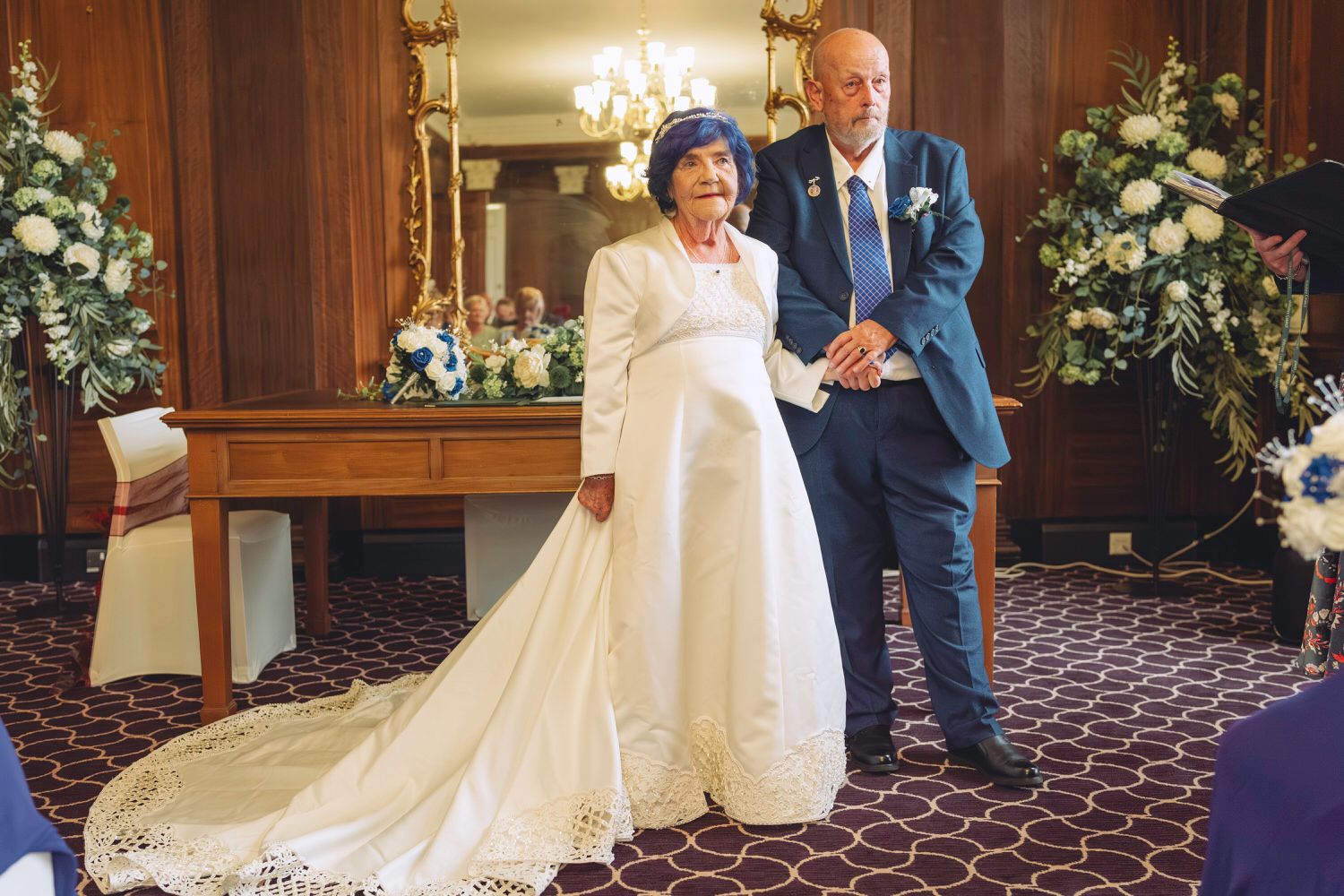 Elegant couple in wedding attire, bride in white gown with train, groom in blue suit, posing in decorated venue.