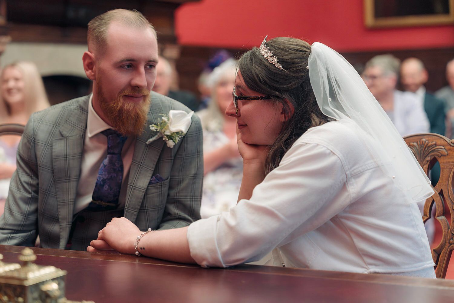 Bride and groom sit together at wedding ceremony table, bride wearing veil and tiara, groom in gray suit with boutonniere.