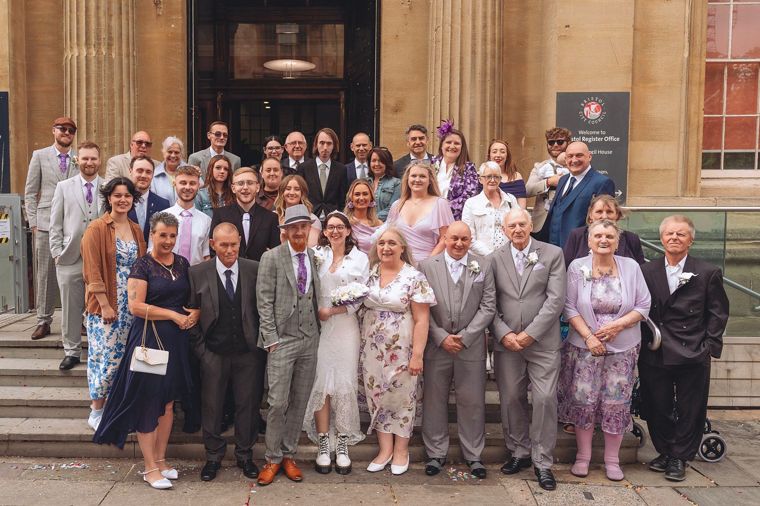 Large wedding party group photo on building steps, bride in white dress with diverse guests.