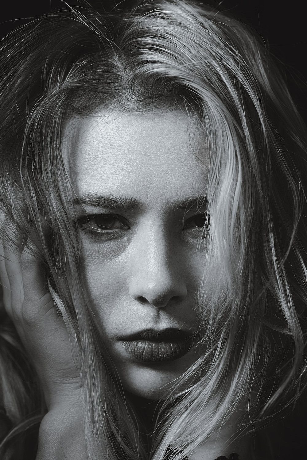 Black and white portrait of a young blonde woman with intense gaze and dark lips, hair framing her face dramatically.