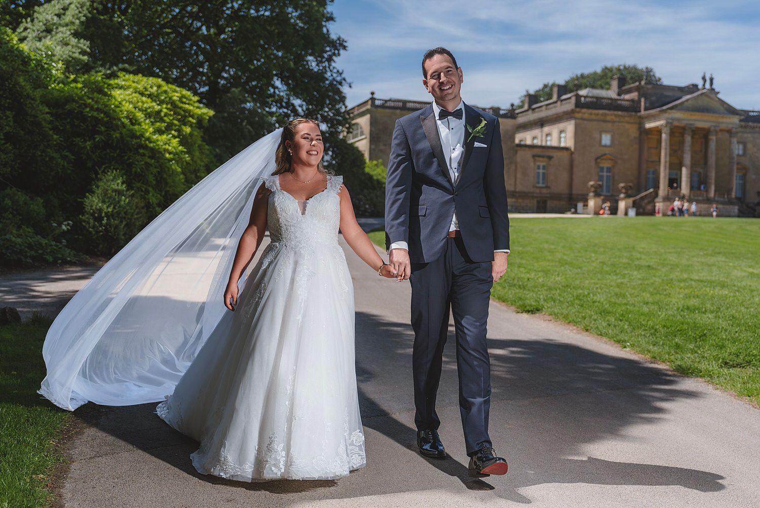 Bride and groom walking hand in hand on a sunny path outside a grand estate on their wedding day.