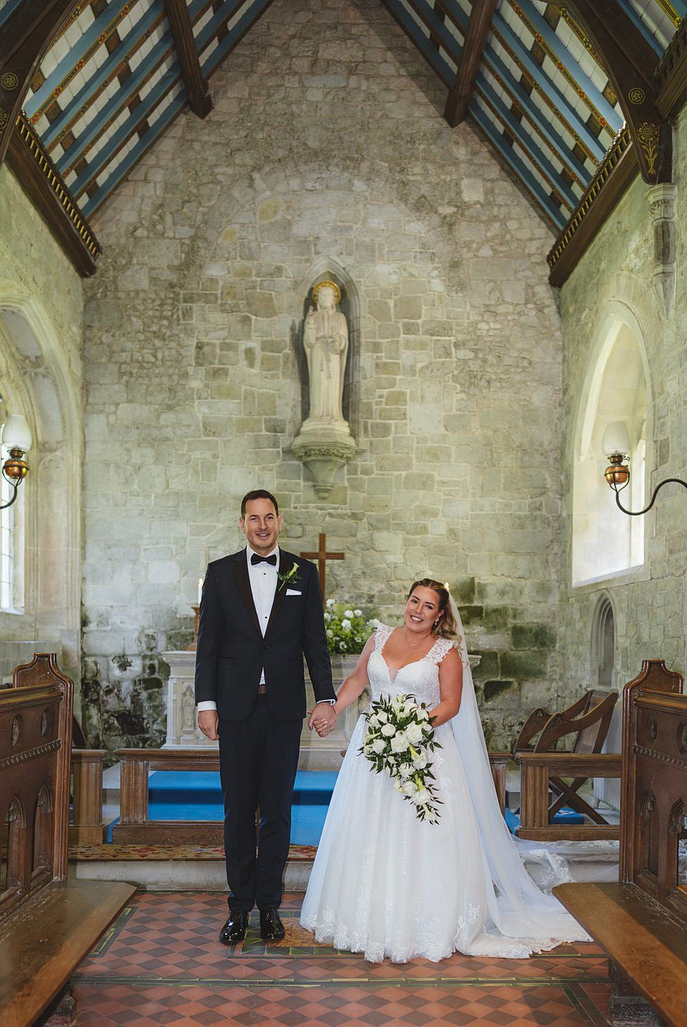 Bride and groom holding hands at church altar, bride in white gown with bouquet, groom in black tuxedo.