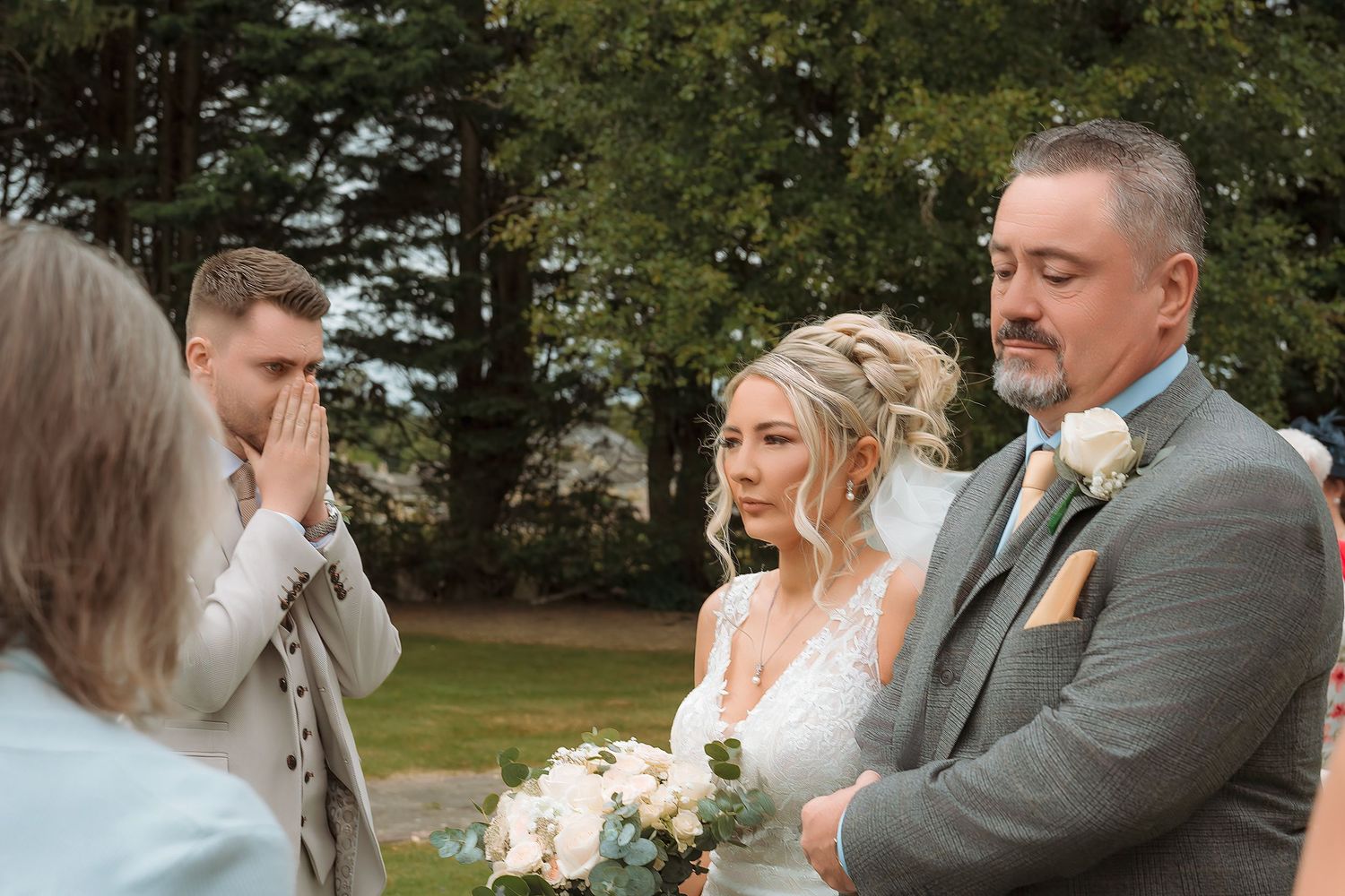 Emotional outdoor wedding ceremony with bride holding bouquet, groom in gray suit, and tearful guest in background.
