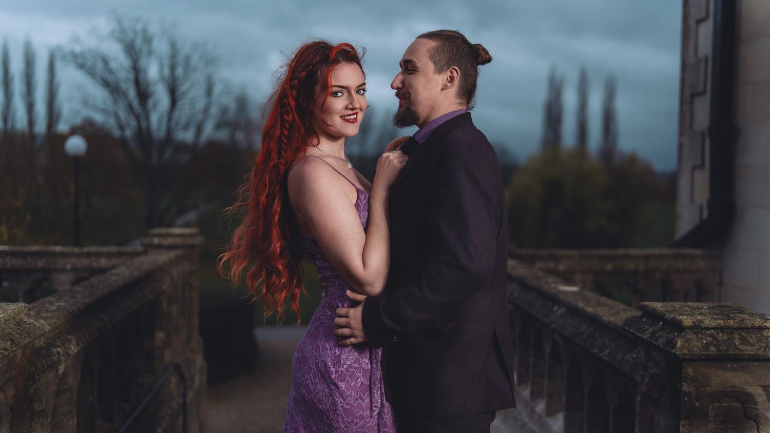 Couple in formal attire posing on stone balcony, woman with red hair in purple dress, man in black suit.