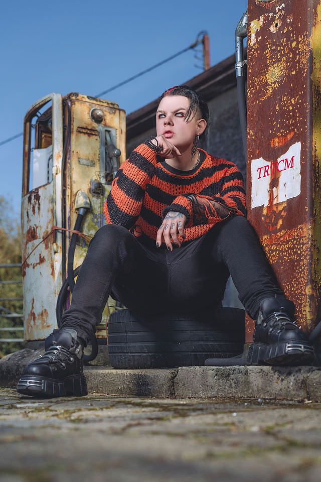 Model in orange striped sweater and platform boots poses by rusty industrial equipment outdoors.