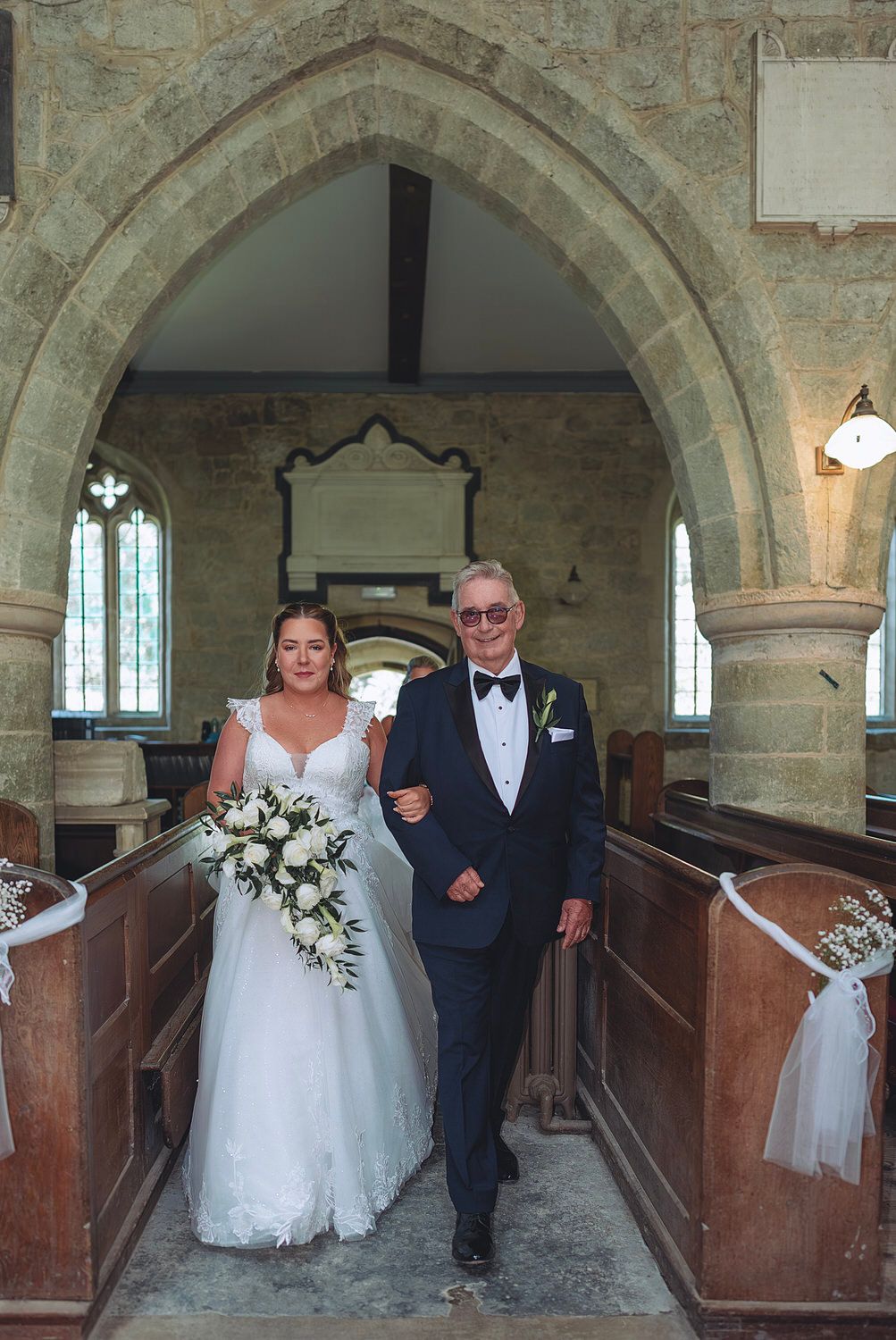Bride in white gown walks down church aisle with older gentleman in tuxedo, holding white floral bouquet.