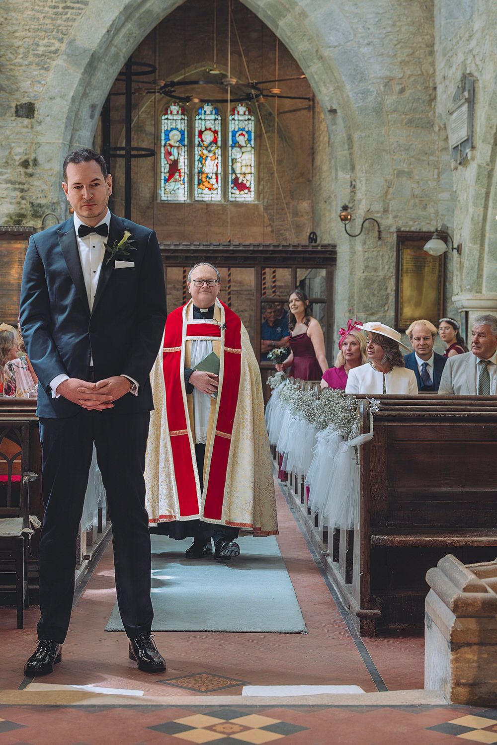Groom in navy tuxedo stands at church altar as officiant walks down aisle with wedding guests seated.