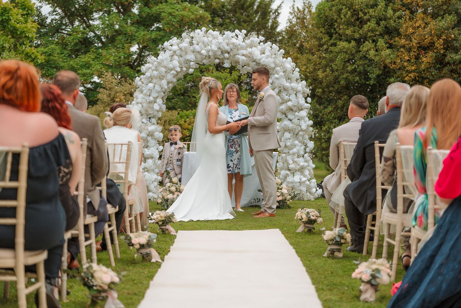 Outdoor wedding ceremony with couple at floral arch altar, white aisle runner, and seated guests on elegant wooden chairs.