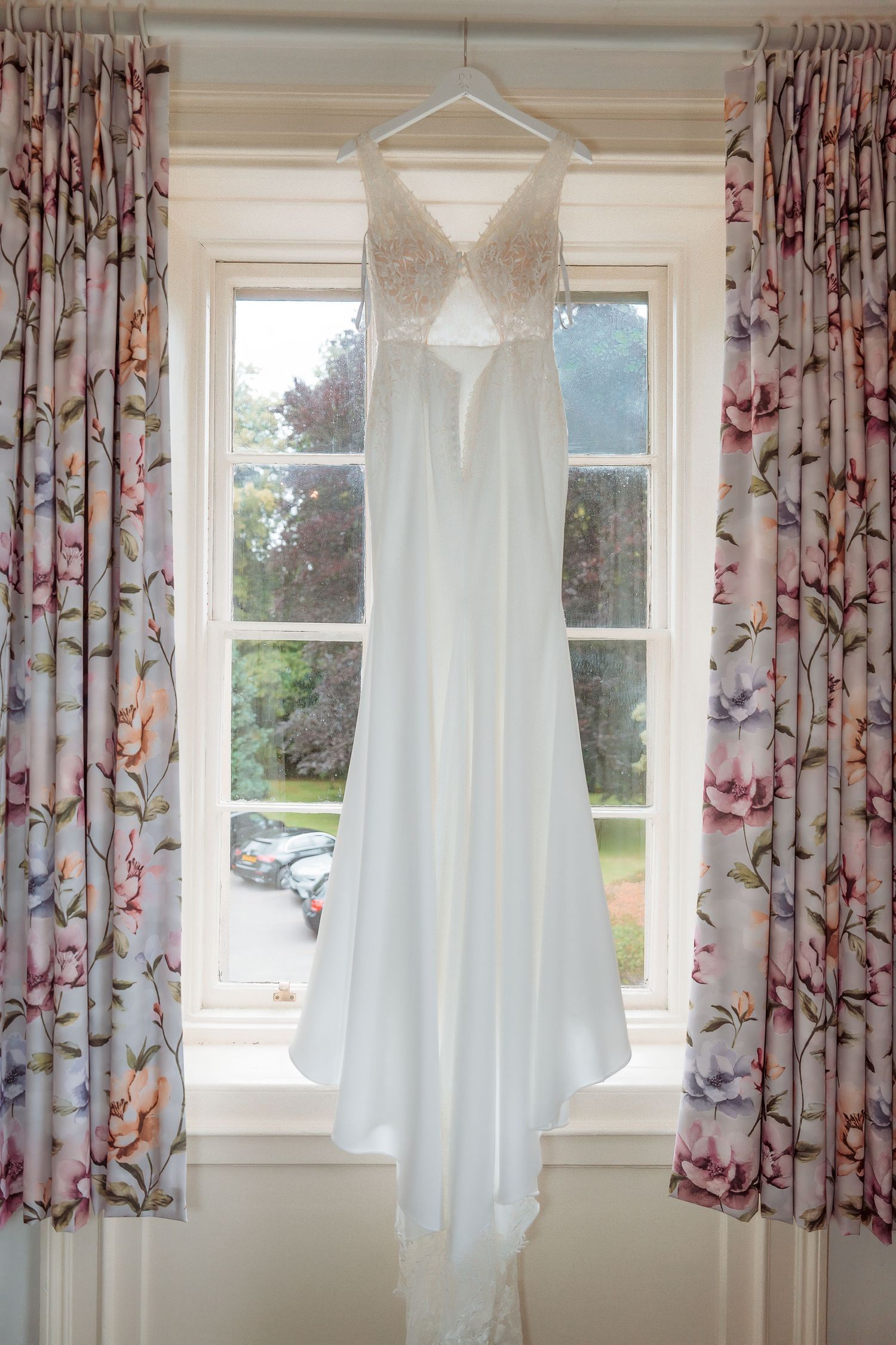White wedding dress with lace bodice hanging on a window with floral curtains, natural light background.