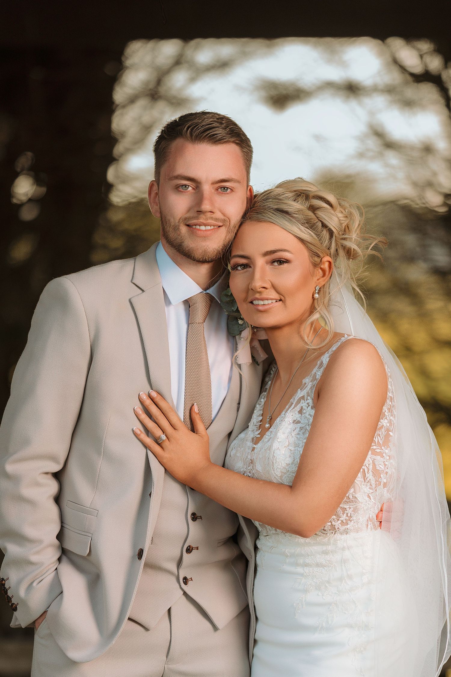 Smiling bride and groom pose together outdoors, she in lace wedding dress with veil, he in beige three-piece suit.