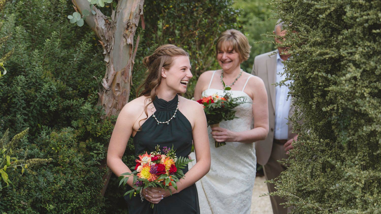 Two bridesmaids in green dresses holding colorful bouquets walk through a lush garden at a wedding ceremony.