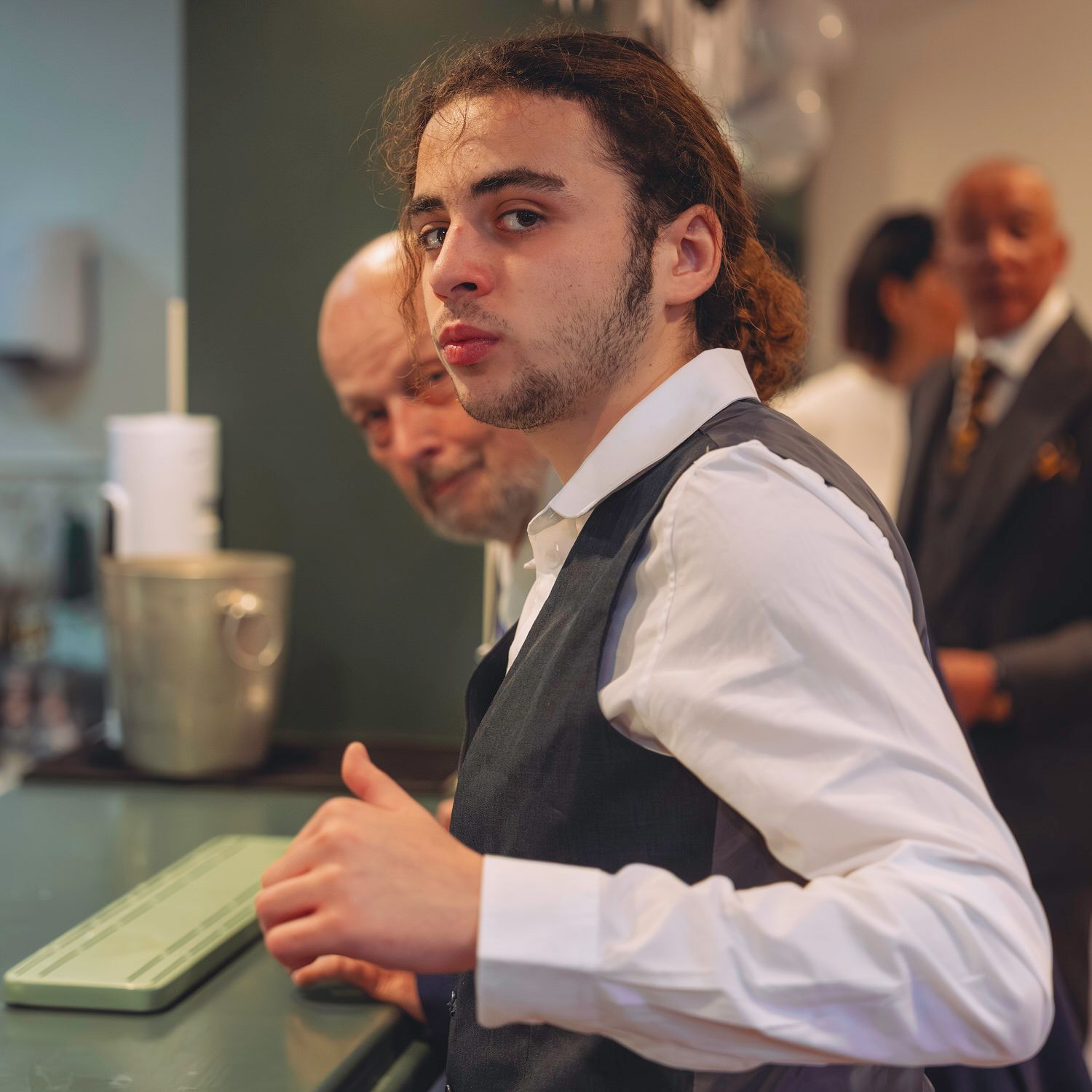 Young man in white shirt and vest working at a counter with a keyboard, with people in the background.