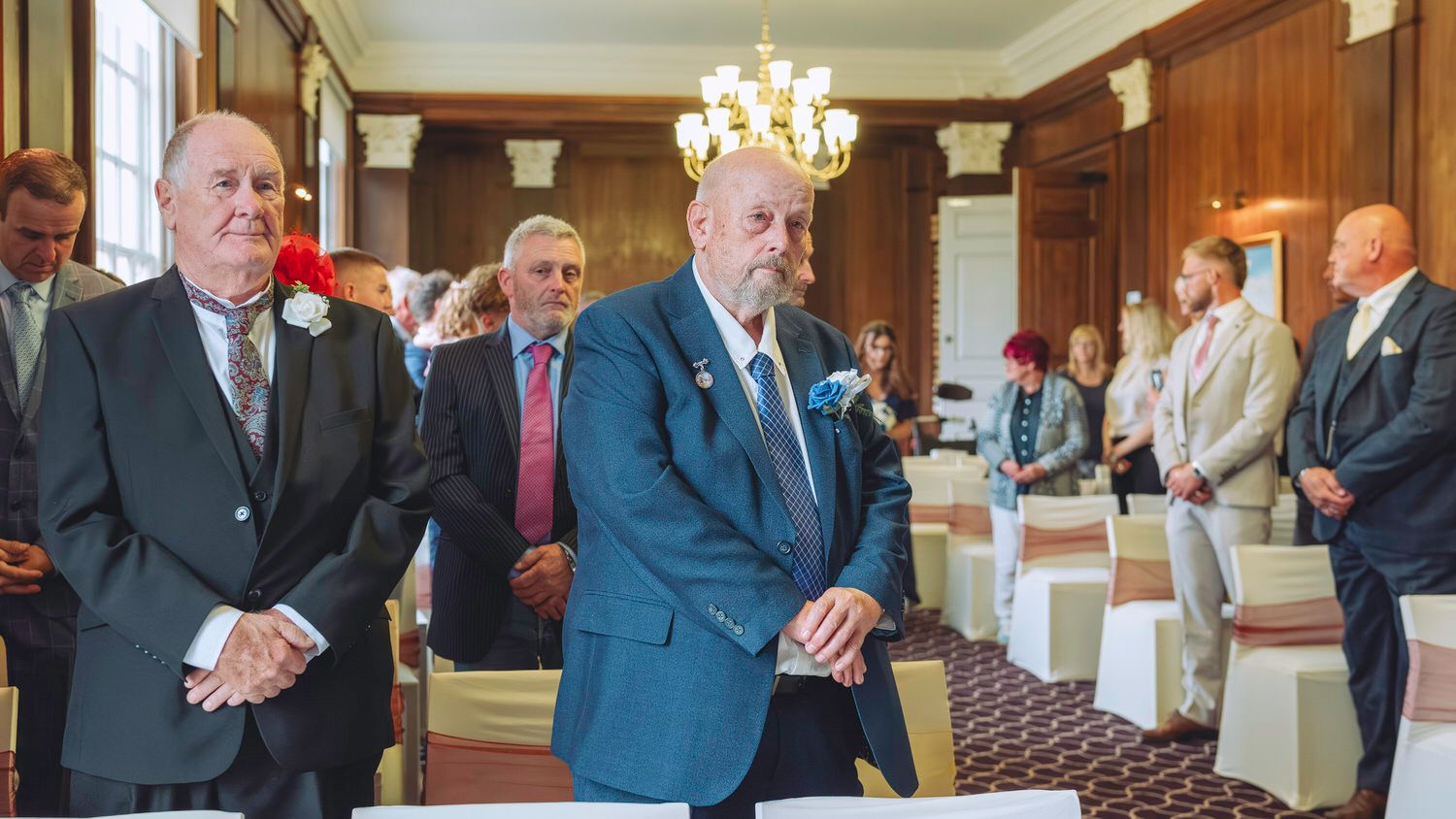 Two men in suits stand at the aisle of an elegant wedding ceremony venue with guests seated in rows.