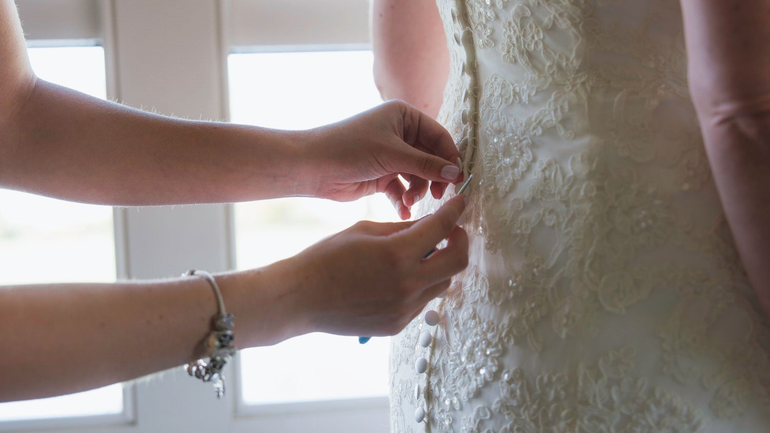 Close-up of hands carefully fastening pearl buttons on the back of a lace bridal gown.