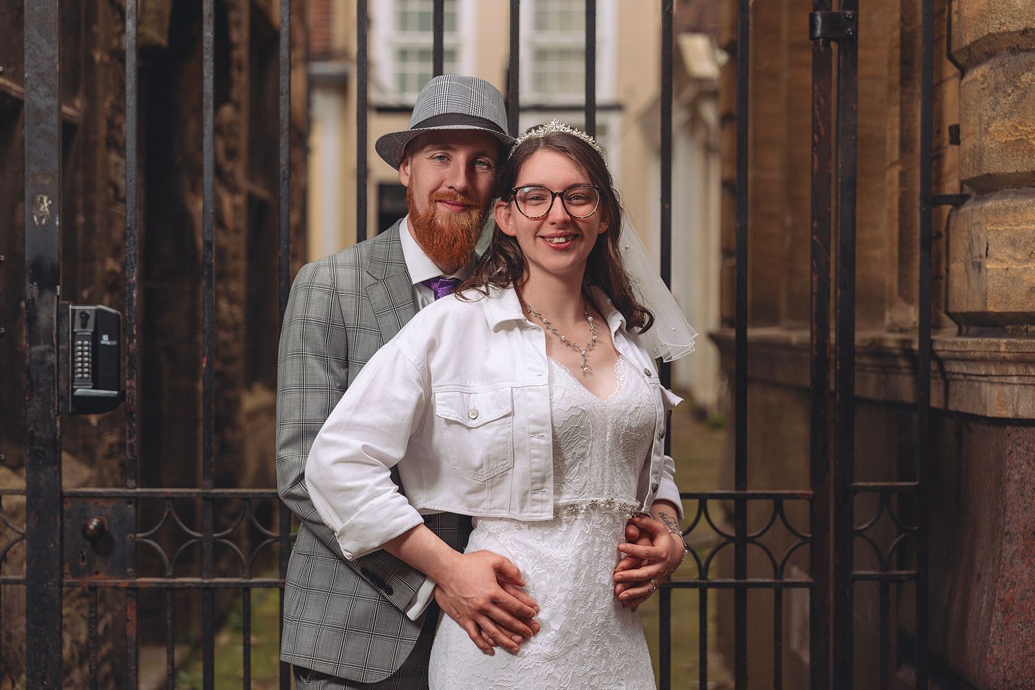 Smiling couple posing by iron gates, groom in grey tweed hat, bride in white dress.