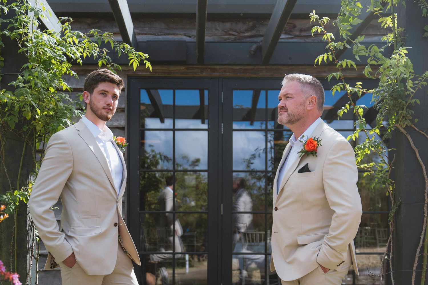 Two grooms in matching tan suits stand outdoors before glass doors, one wearing an orange boutonniere at a wedding.