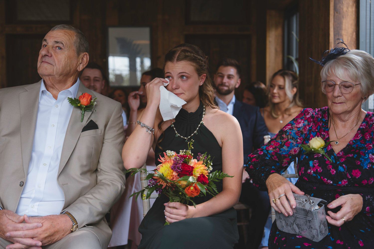 Emotional bridesmaid wiping tears at wedding ceremony while holding colorful bouquet with guests seated nearby.