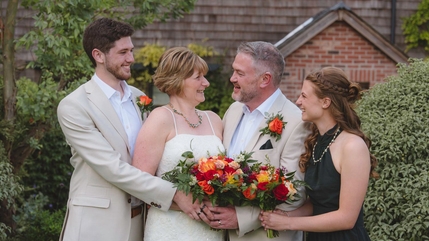 Wedding couple with bouquet pose alongside two guests in a garden setting near a brick building.