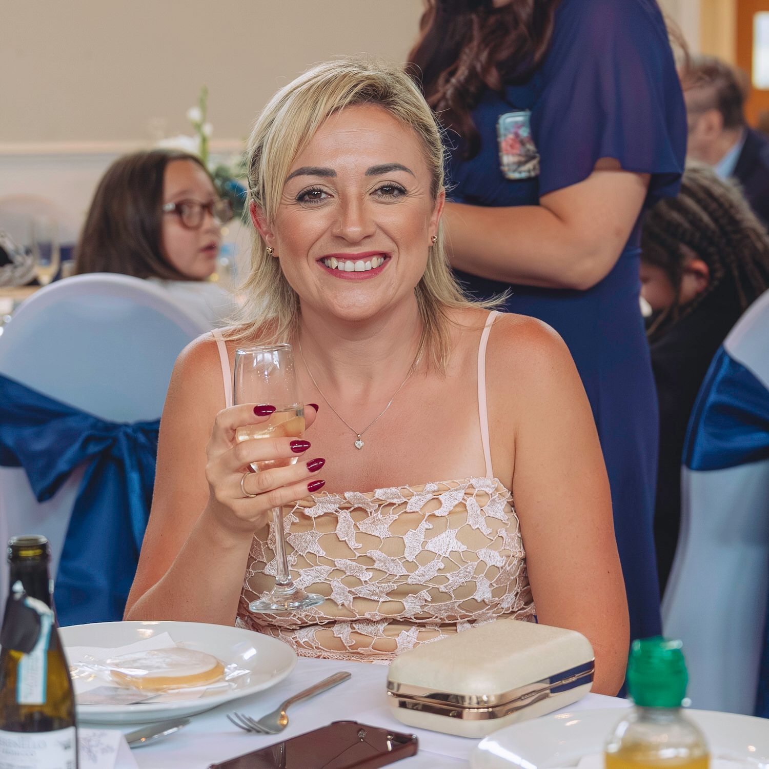 A smiling blonde woman in a floral dress holds a drink at an elegant wedding reception with blue satin chair bows.