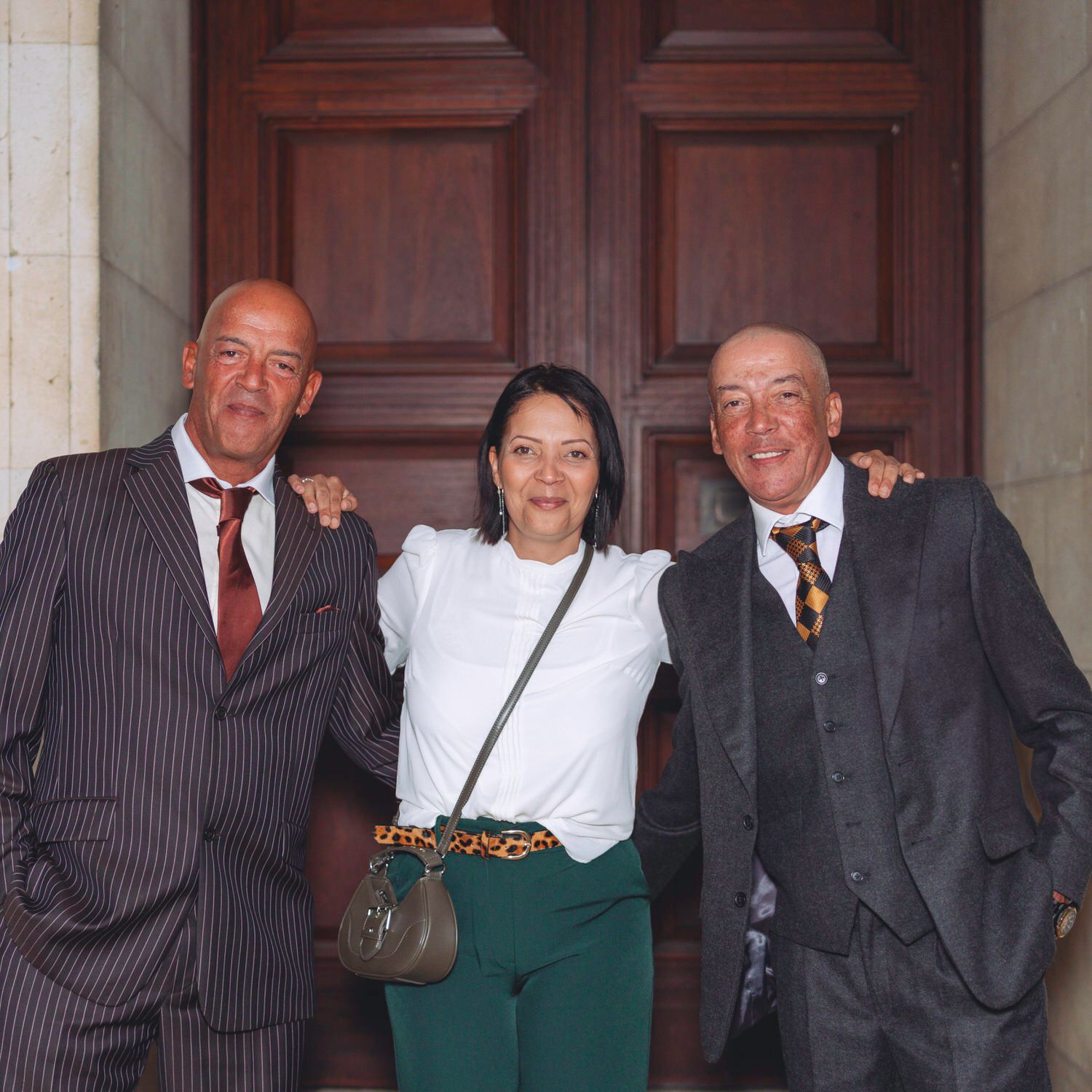 Three elegantly dressed people posing together in front of large wooden doors.