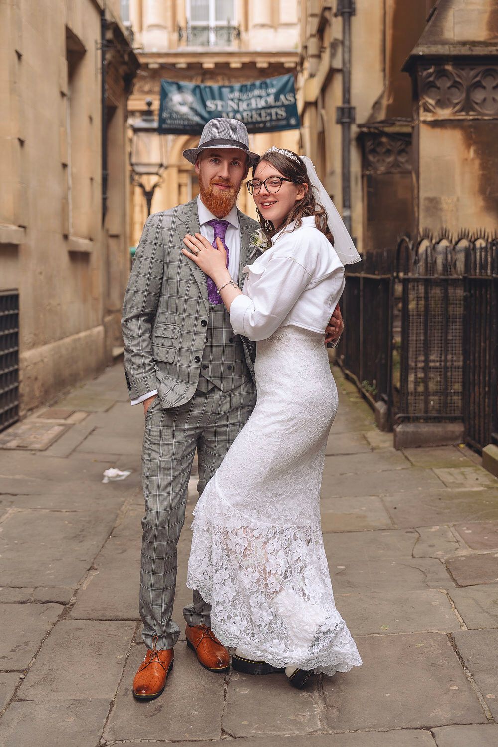 A groom in a plaid suit and hat stands with his bride in a white lace dress in a historic cobblestone alley.