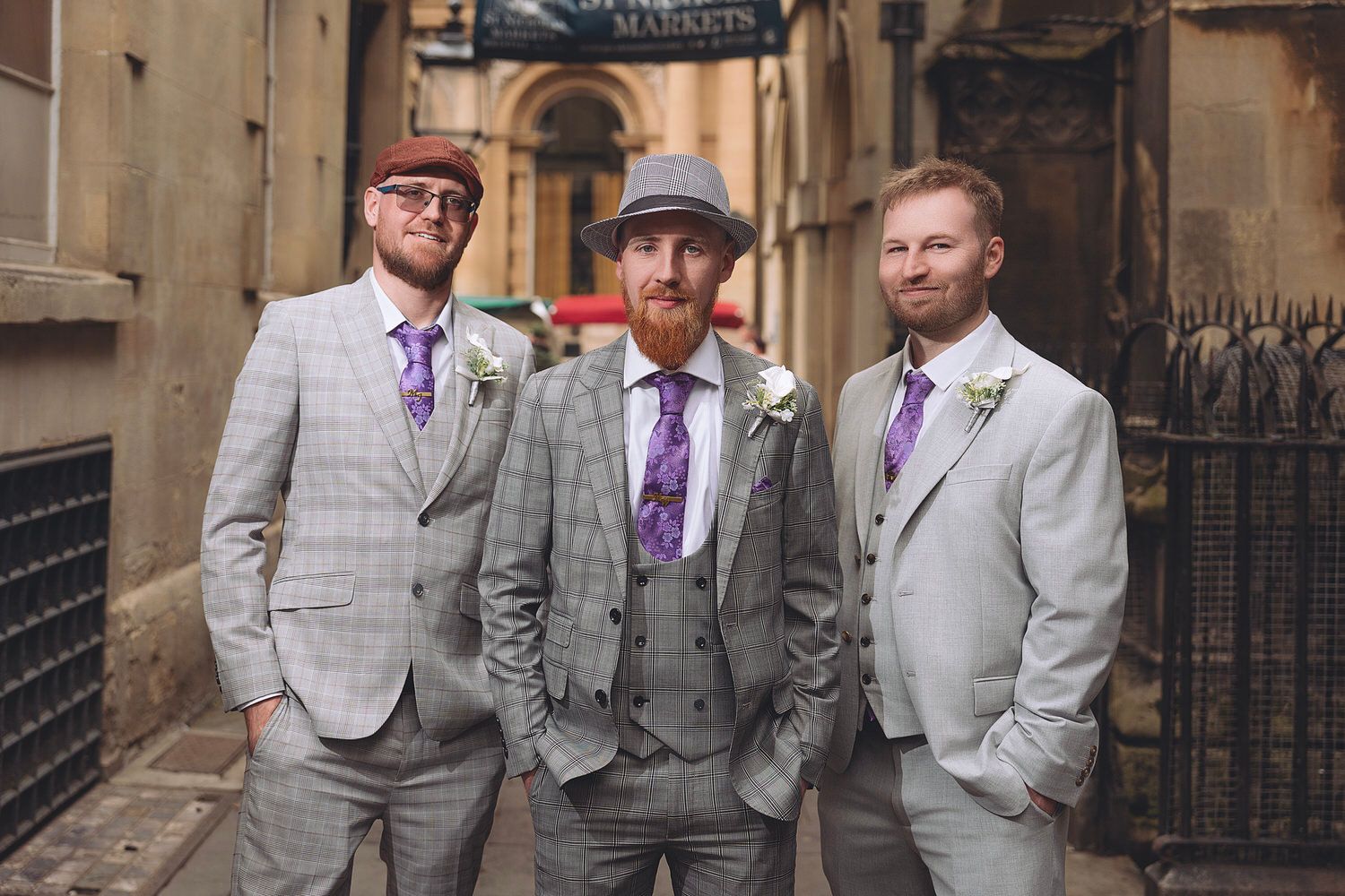 Three groomsmen in matching grey plaid suits with purple ties pose in a historic cobblestone alley.