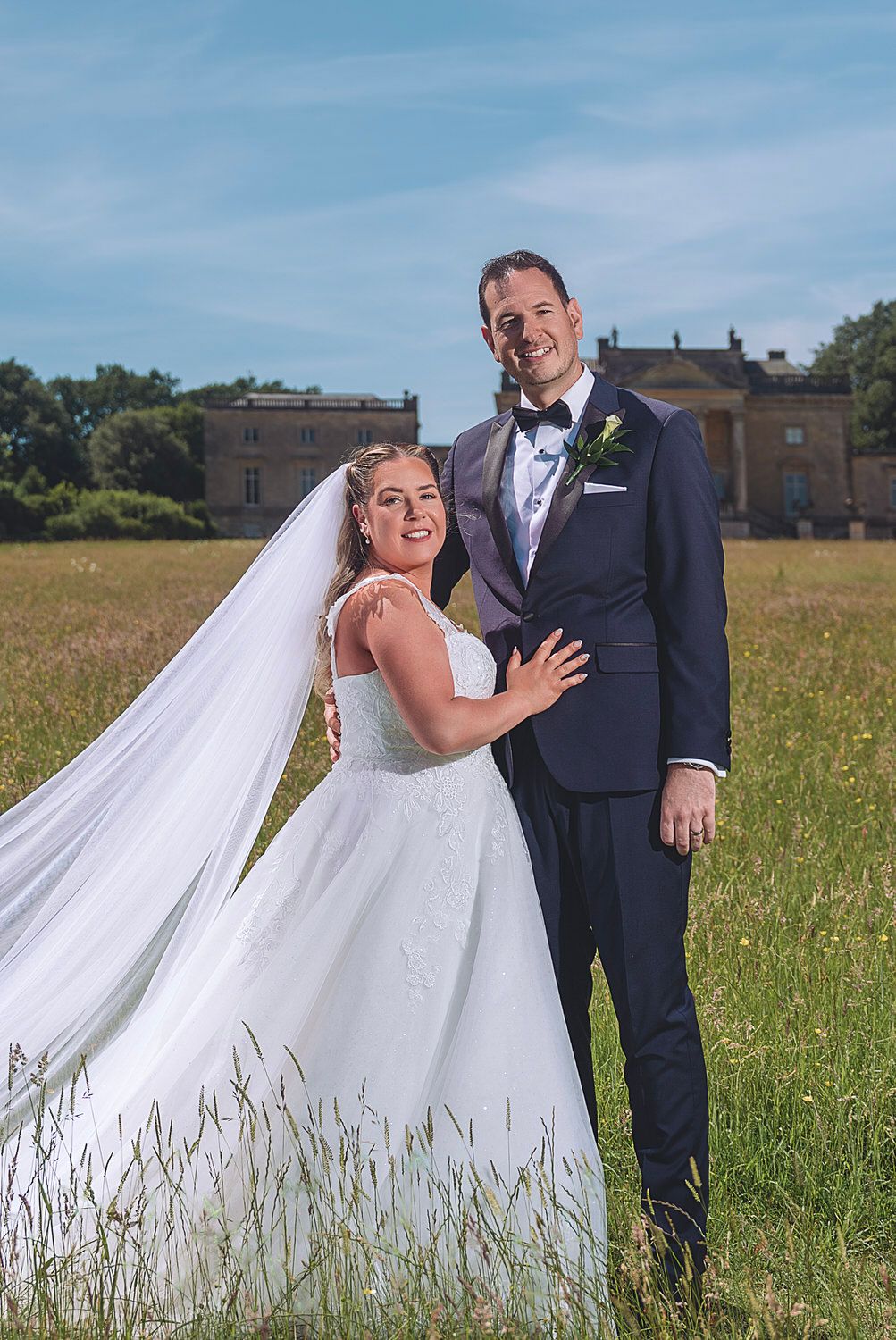 Bride and groom posing in a sunny meadow with a grand estate in the background on their wedding day.