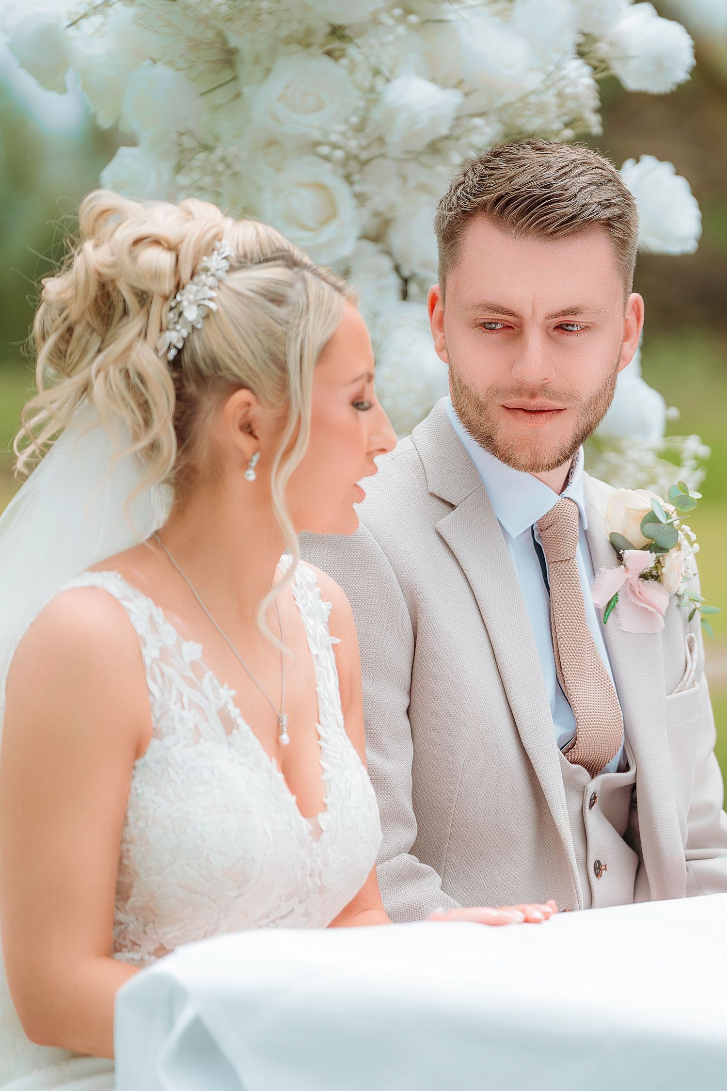 Bride in white lace gown and groom in beige suit share an intimate moment at outdoor wedding ceremony with white floral arch.