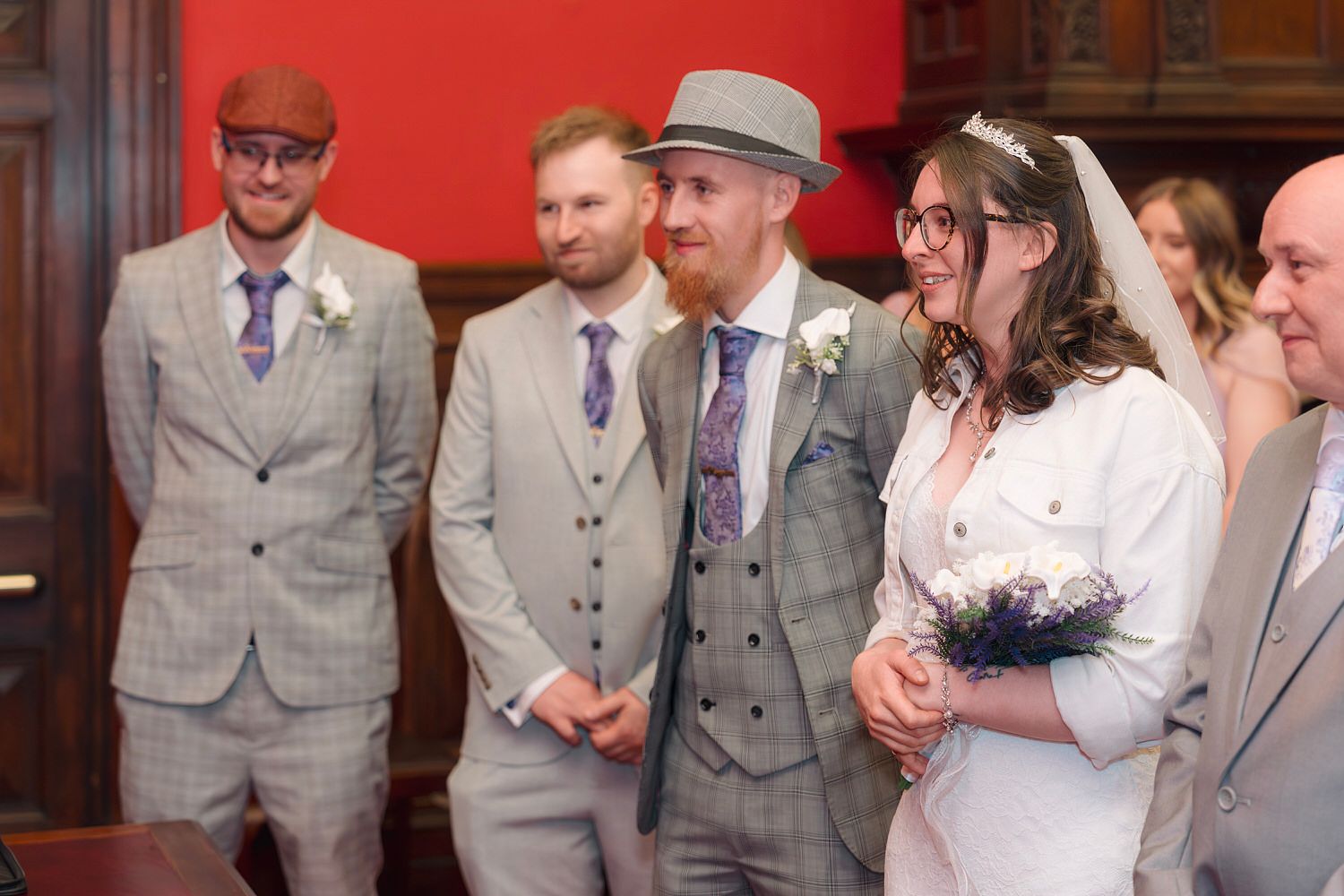 Bride and groom smiling during wedding ceremony, groomsmen in grey checked suits standing behind them.