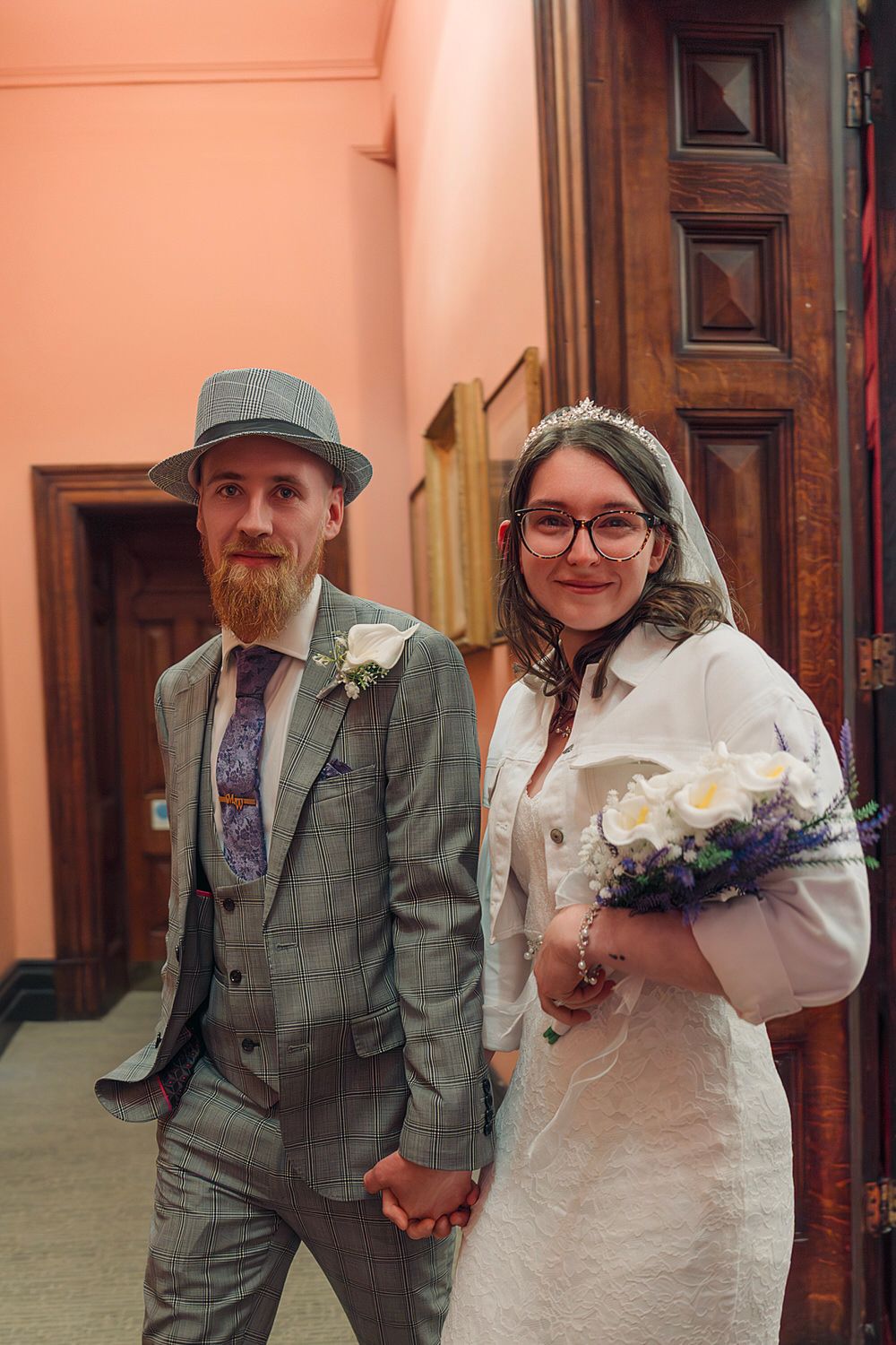 A bearded groom in a gray suit and hat holds hands with a bride in a white dress holding a bouquet.