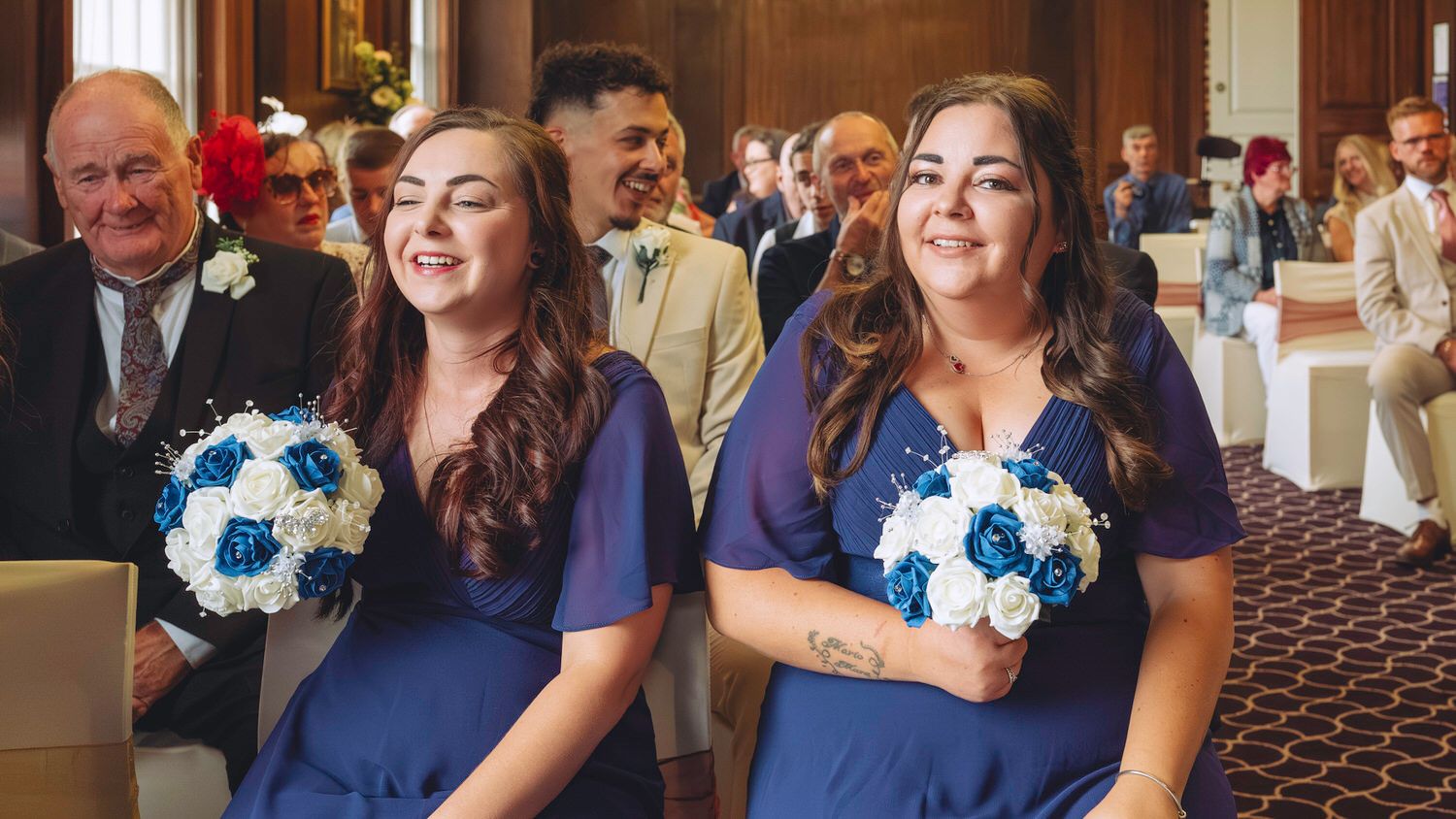 Two bridesmaids in navy blue dresses holding blue and white bouquets smile during a wedding ceremony.