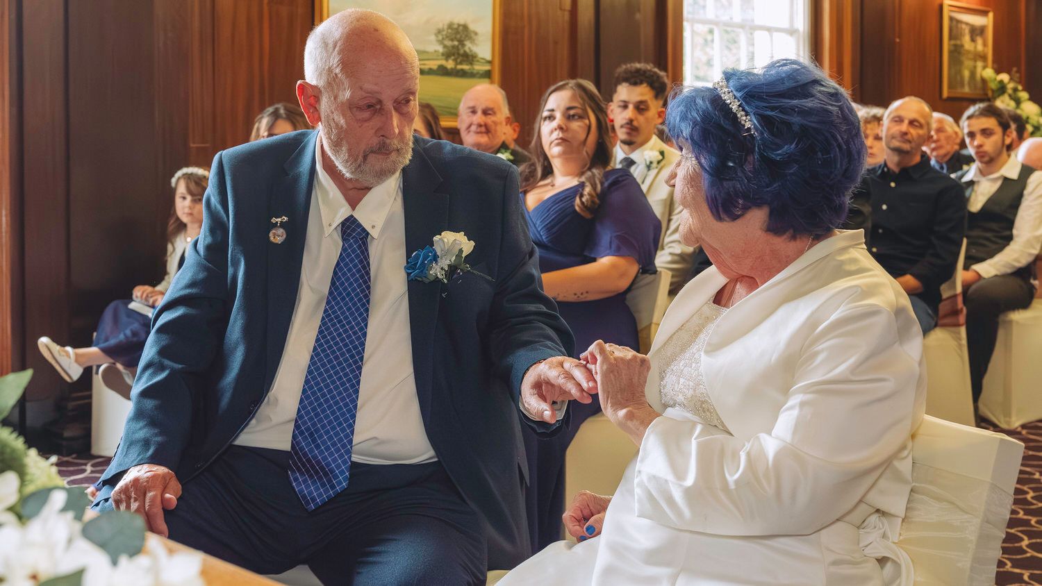 Elderly couple exchanging rings at a wedding ceremony, groom in blue suit, bride in white outfit with blue hat.