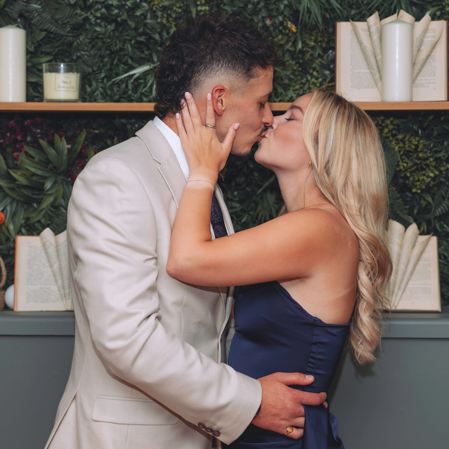 Couple sharing a romantic kiss in front of a green botanical wall with candles and books on shelves.