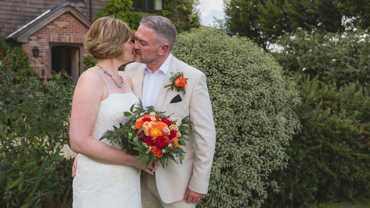 Bride and groom share a romantic kiss outdoors, bride holding vibrant orange bouquet, groom in tan suit.