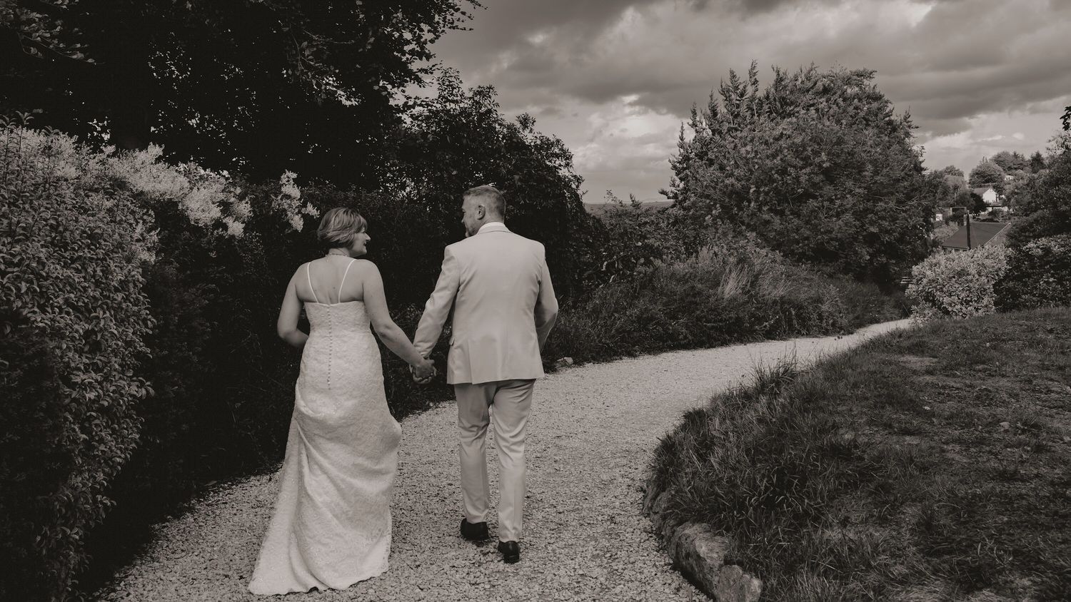 Bride and groom walk hand in hand down a winding garden path in black and white.
