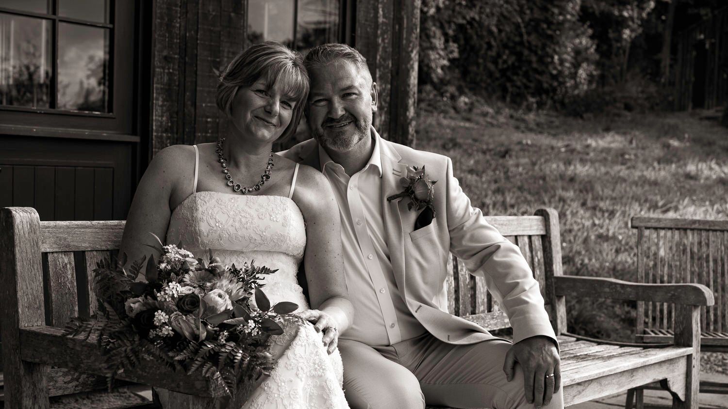 Black and white wedding photo of smiling bride and groom sitting on a bench outdoors holding a bouquet.