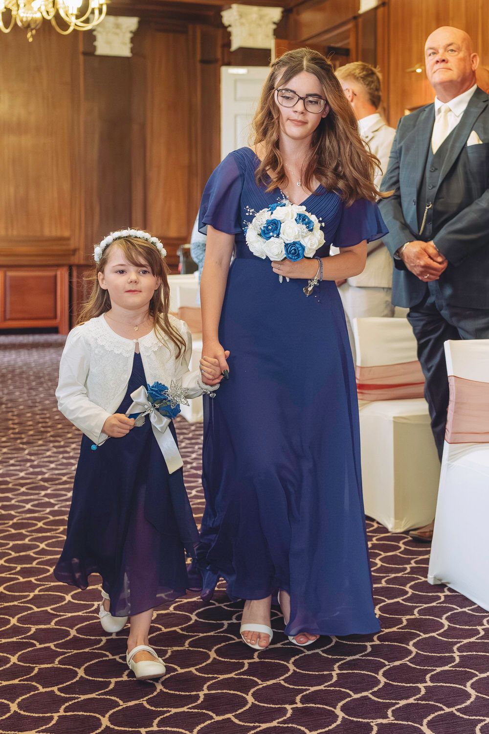 A bridesmaid in navy blue gown holds bouquet walking with young flower girl in matching dress at wedding ceremony.