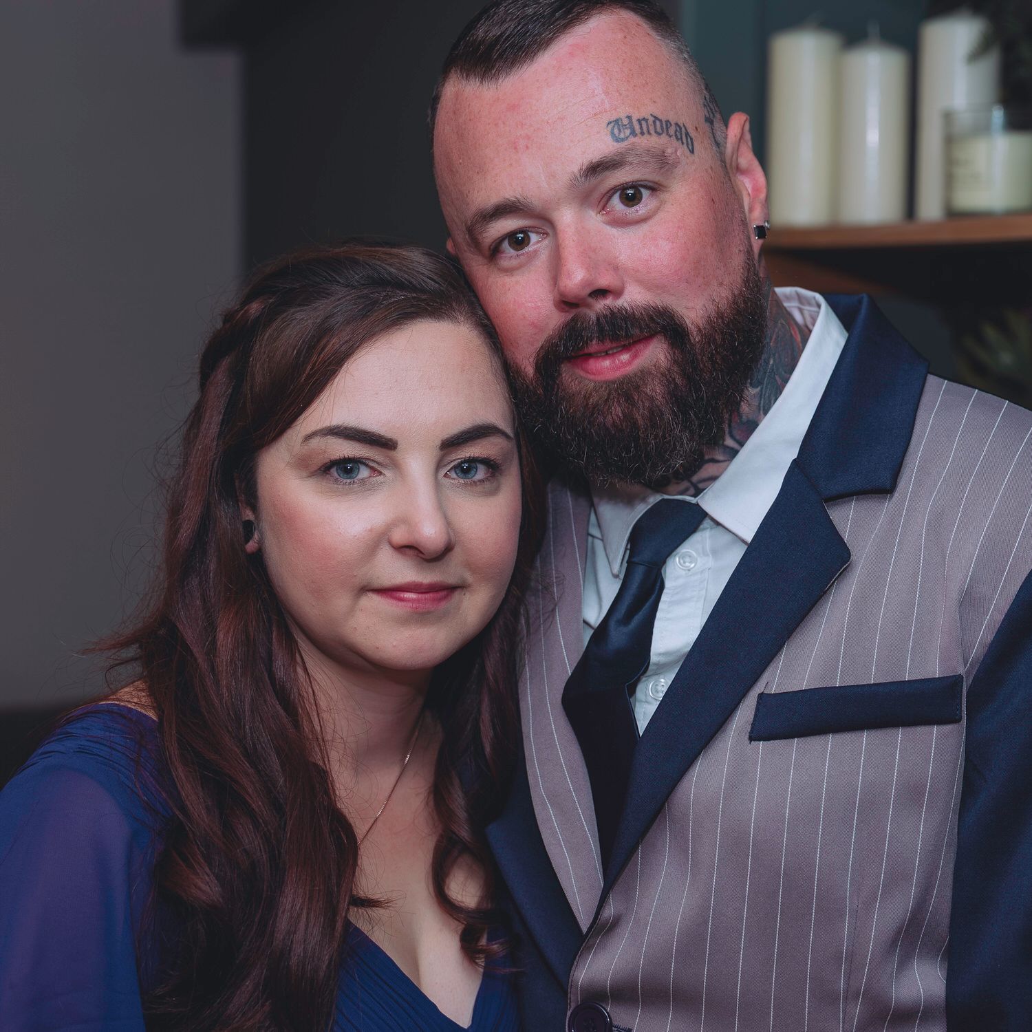 A couple poses closely together, the woman in blue and the man in a pinstripe vest with a neck tattoo.