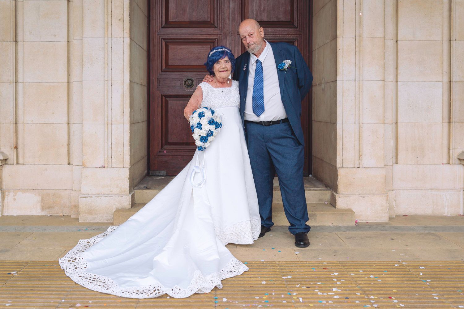 Bride in white gown with blue bouquet and groom in navy suit pose outside church doors on wedding day.
