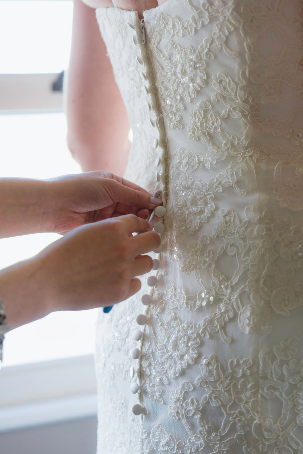 Hands buttoning the back of a white lace wedding dress with elegant floral detailing.