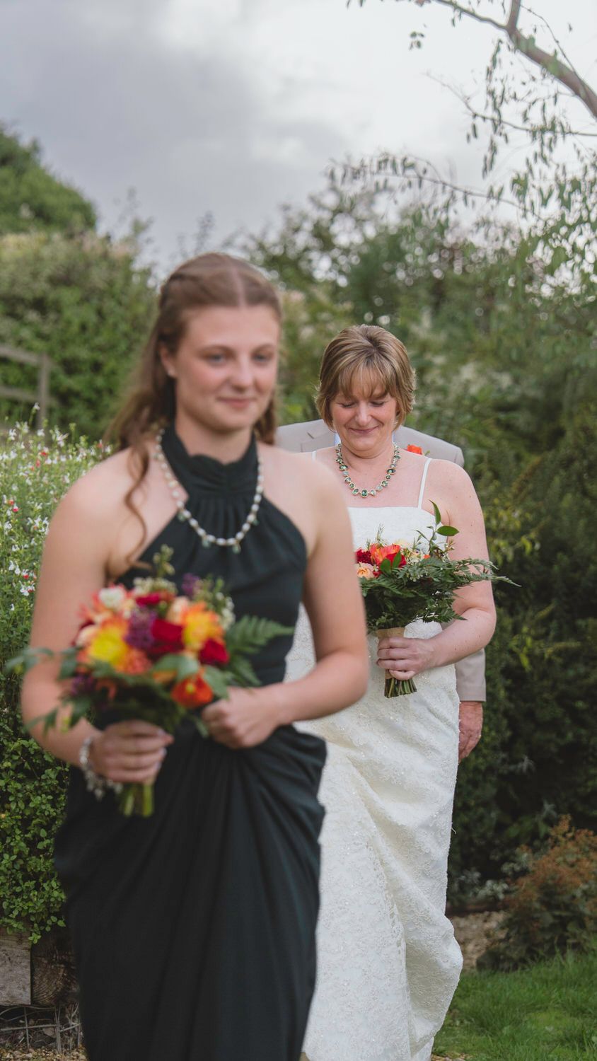 Bridesmaid in black dress leads bride outdoors carrying vibrant bouquets at garden wedding ceremony.