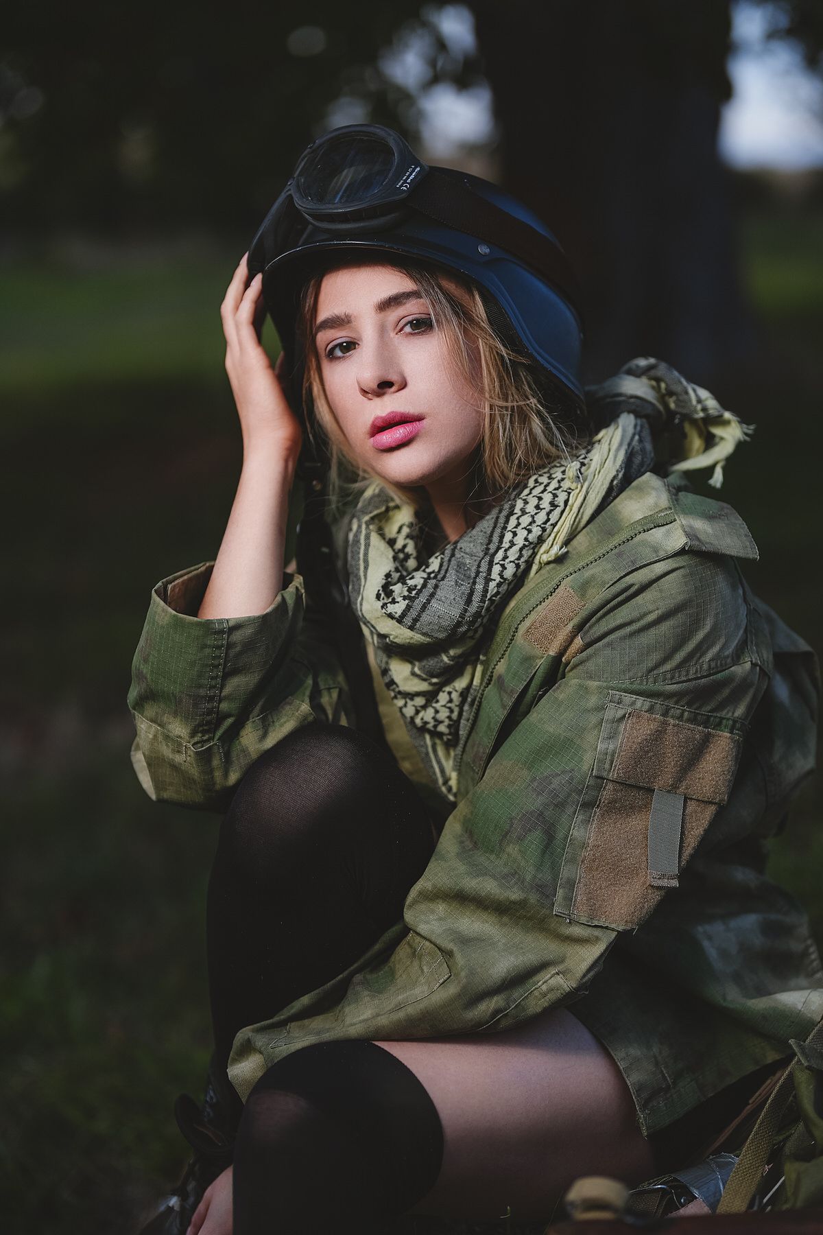 Young woman in military camouflage jacket and black helmet with goggles posing outdoors.