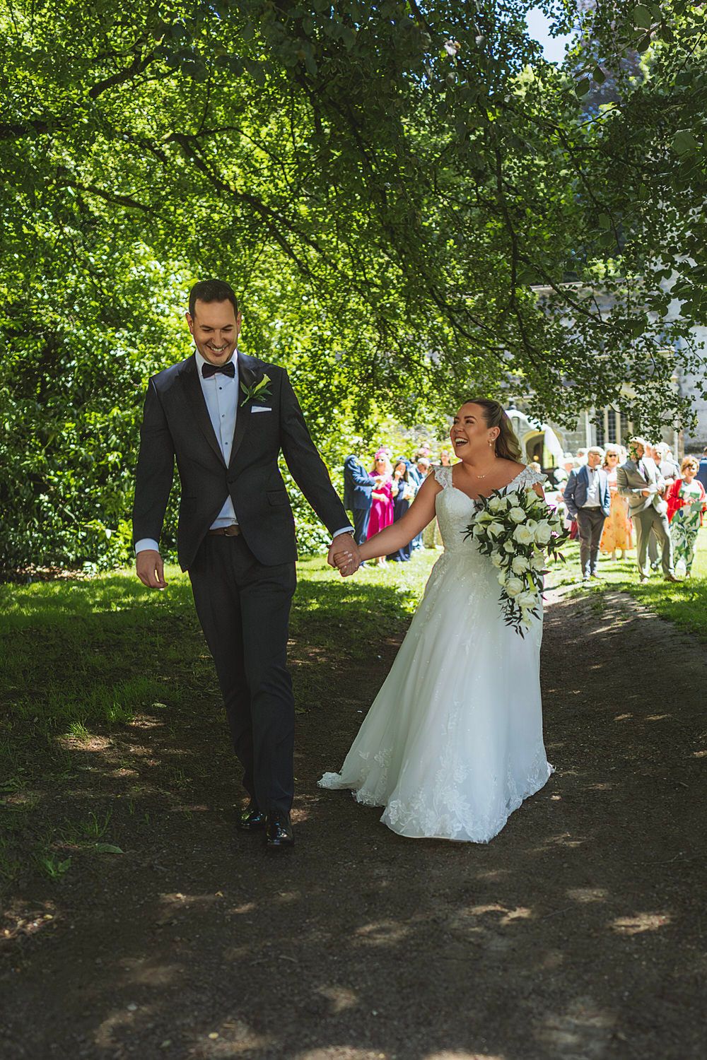 Newlywed couple holding hands walking on garden path, bride in white gown with bouquet, groom in dark tuxedo.