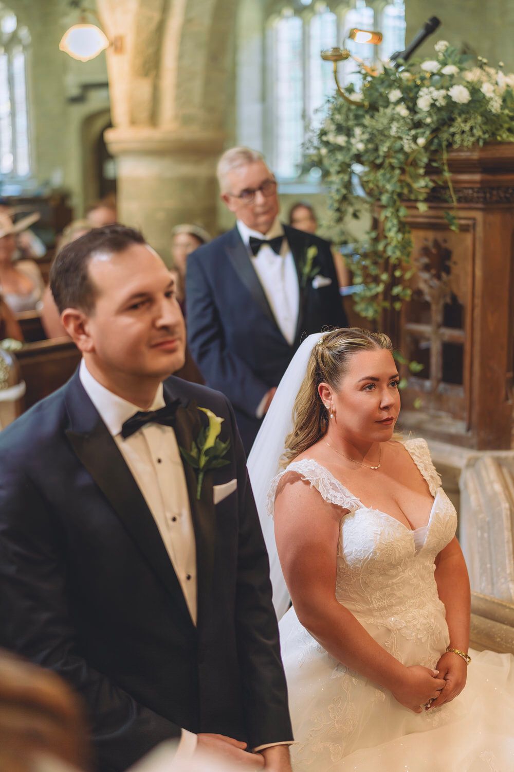 Bride and groom stand at church altar during wedding ceremony, bride in lace gown with veil, groom in navy tuxedo.