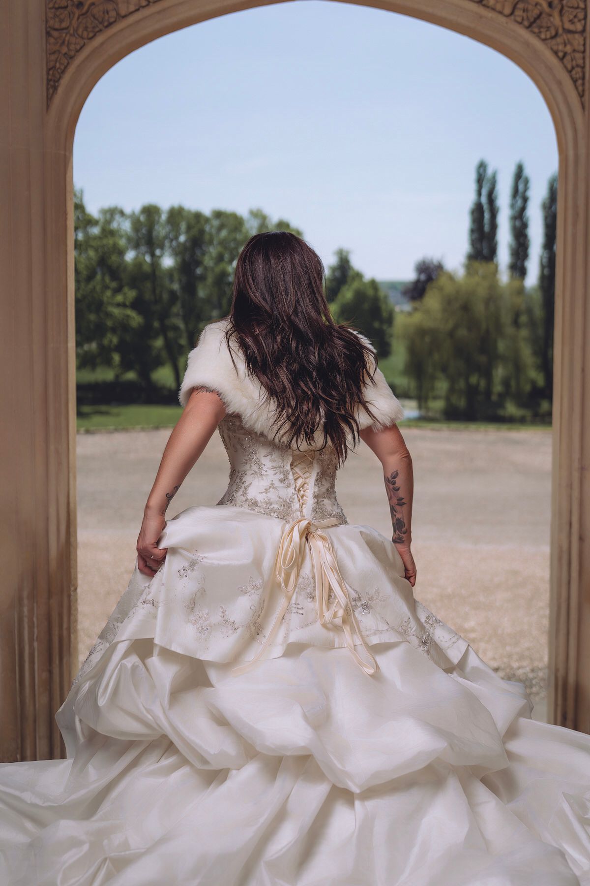 Bride in elegant layered white gown with fur shrug stands in archway overlooking lush green garden.