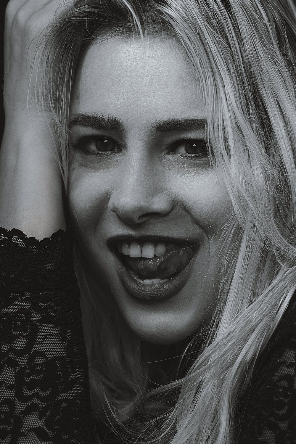 Black and white portrait of a smiling blonde woman with an open mouth, wearing lace top, hand near face.