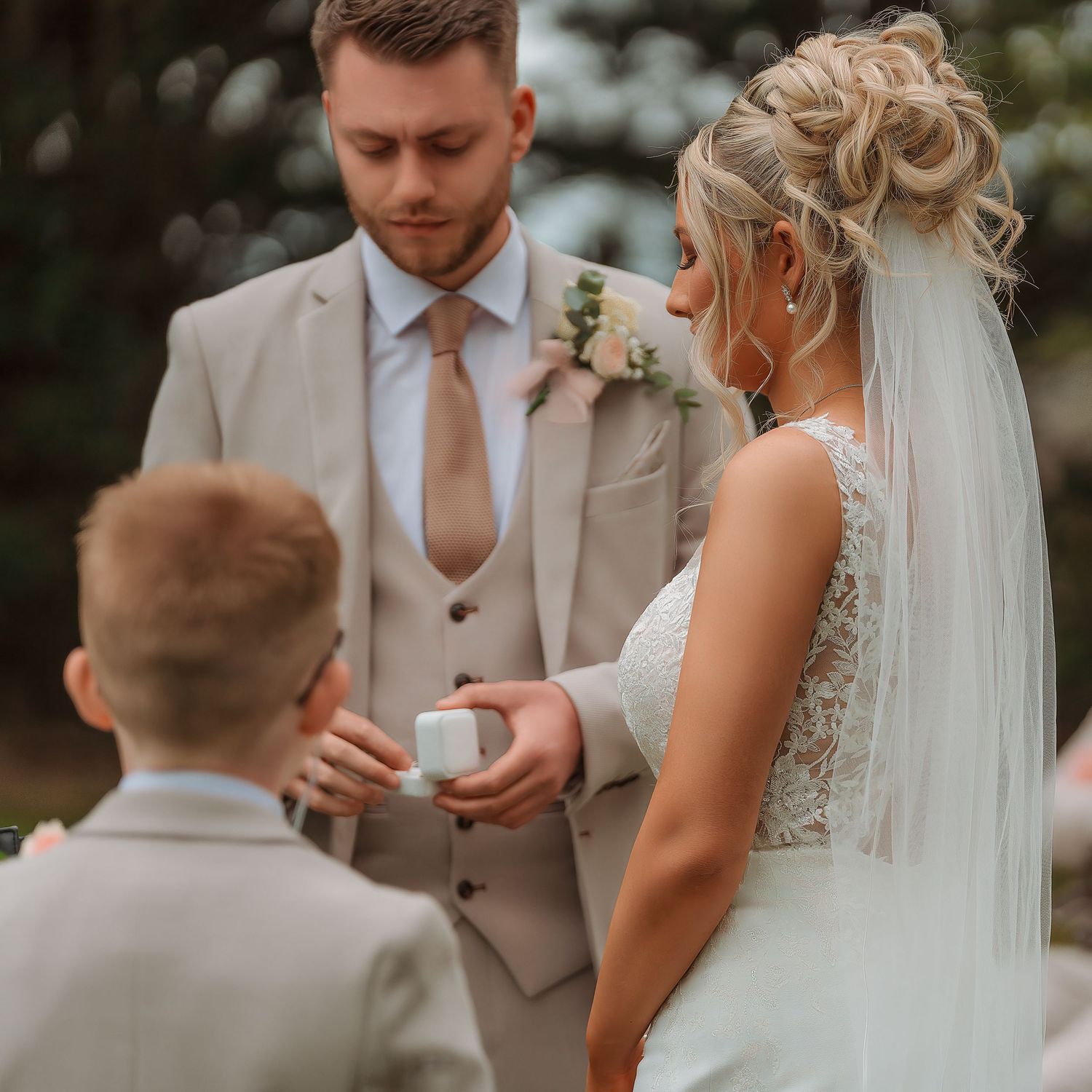 A groom in a beige suit and bride in a white lace dress exchange rings with a young boy at an outdoor wedding ceremony.