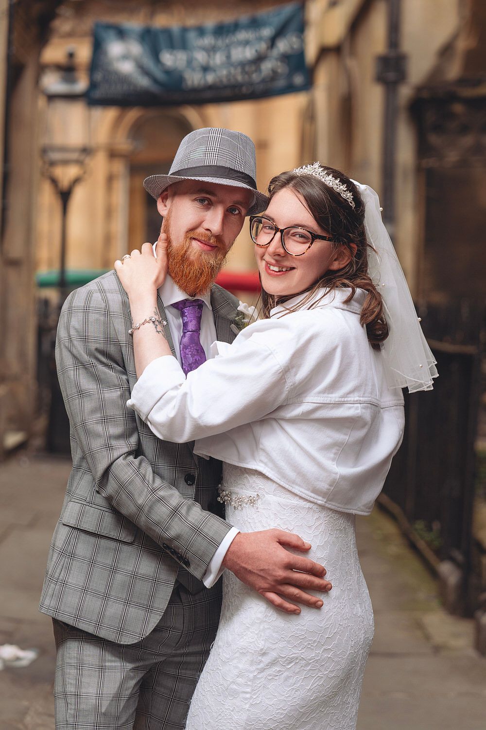 A smiling bride and groom in wedding attire embrace outdoors, he wears a grey suit and hat, she wears a white lace dress.